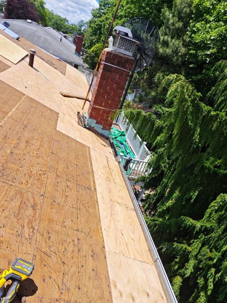 View of a house roof under repair in NY, with plywood sheets partially installed. A chimney, satellite dish, and tools are visible. Trees and rooftops fill the sunny background—a scene familiar to any bathroom remodeler Nassau County trusts.