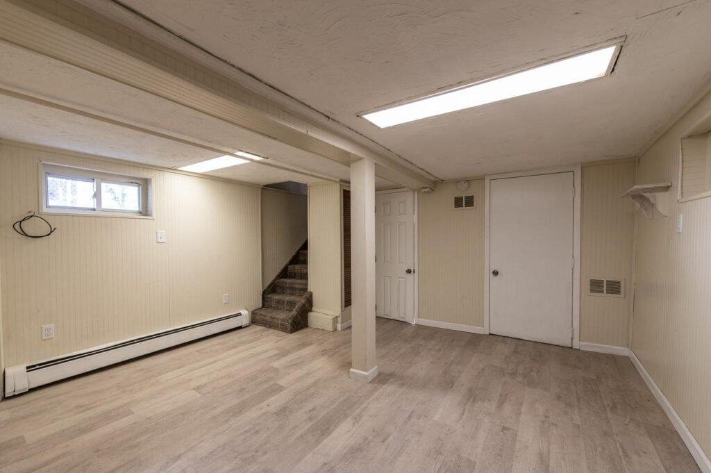 A finished basement room with light-colored walls and wood-style flooring, completed by a skilled general contractor Nassau County, NY. Features recessed lighting, a small window, carpeted staircase, and multiple closed white doors.