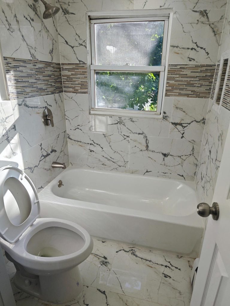 A small bathroom with marble-patterned tiles, a white bathtub, a toilet with the lid up, and a sunlit window above the tub features a band of mosaic tiles—expertly finished by a general contractor in Nassau County, NY.