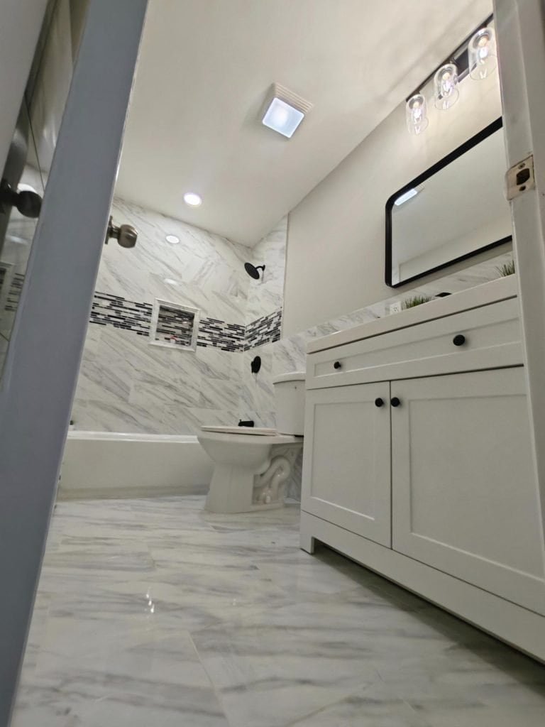 Modern bathroom in NY with marble tile flooring and walls, a white vanity with black-framed mirror, toilet, and bathtub featuring a mosaic tile accent. Bright lighting and clean design by a top bathroom remodeler Nassau County.