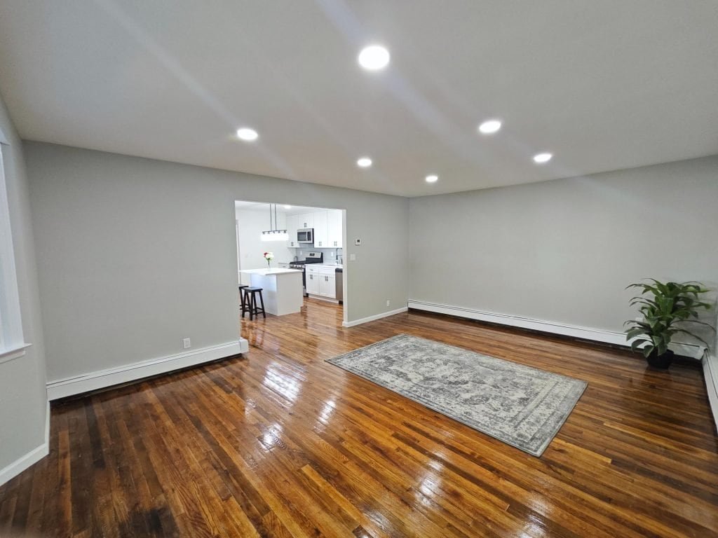 A spacious living room with polished wooden floors, a patterned rug, and neutral-colored walls flows into a bright modern kitchen, thoughtfully designed by a top kitchen remodeler in Nassau County, NY.