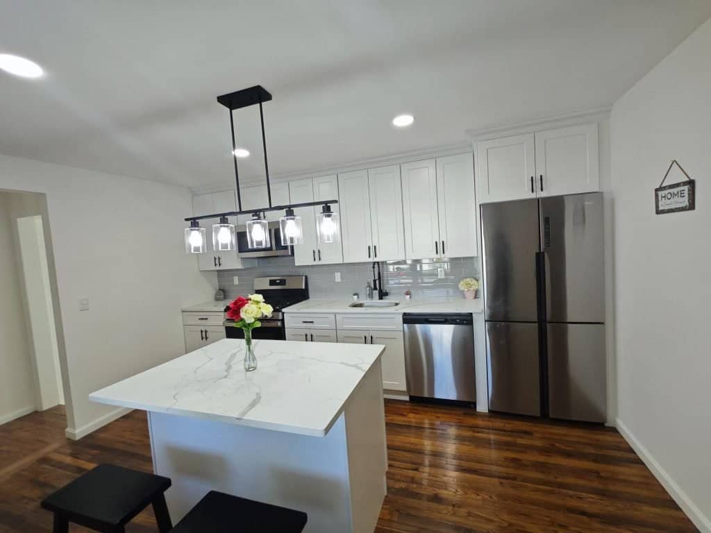 Modern kitchen in NY with white cabinets, stainless steel appliances, a marble kitchen island, black barstools, wood flooring, and bright lighting. Designed by a general contractor Nassau County for a clean, minimalist look.