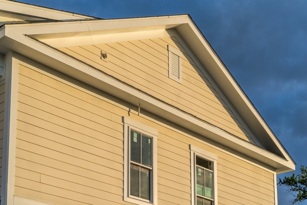 Close-up view of the side of a beige house with horizontal vinyl siding, featuring two white-trimmed windows and a triangular gable roof, set against a partly cloudy sky with patches of blue visible through the clouds.