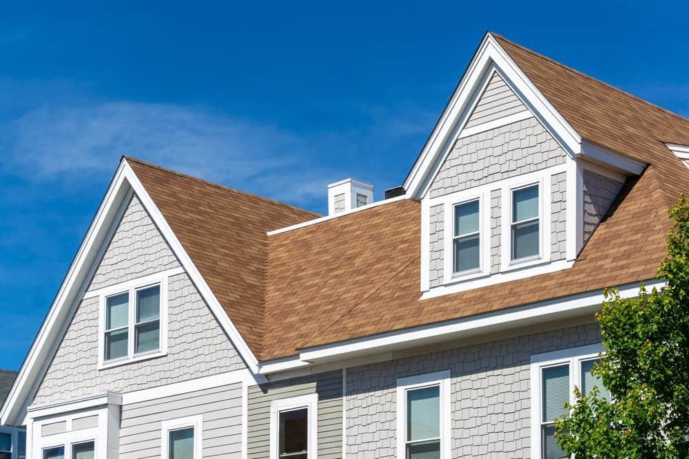 A close-up view of a modern house with light gray siding, white trim, and a brown shingle roof against a clear blue sky.