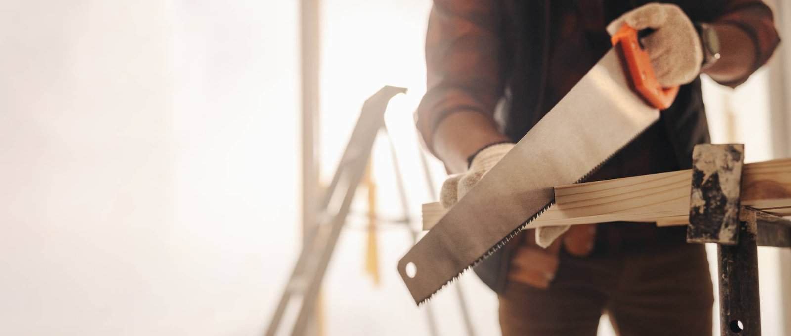 A person wearing gloves uses a handsaw to cut a wooden plank on a workbench, with a blurred background showing a ladder and bright natural light.