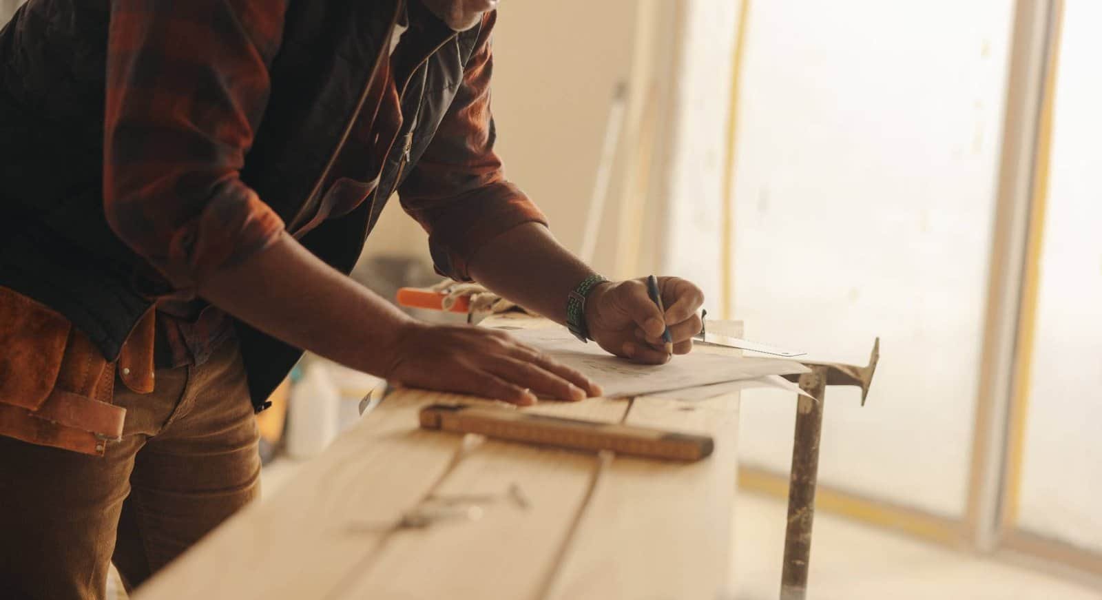 A person wearing a plaid shirt and tool belt is leaning over a wooden plank, using a pencil to mark measurements on paper. Tools are scattered on the workbench in a well-lit workshop.