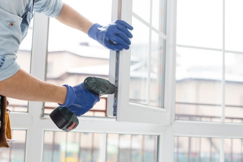 A person wearing blue gloves uses a cordless power drill to install or repair a white-framed window in a bright room with large glass panes.
