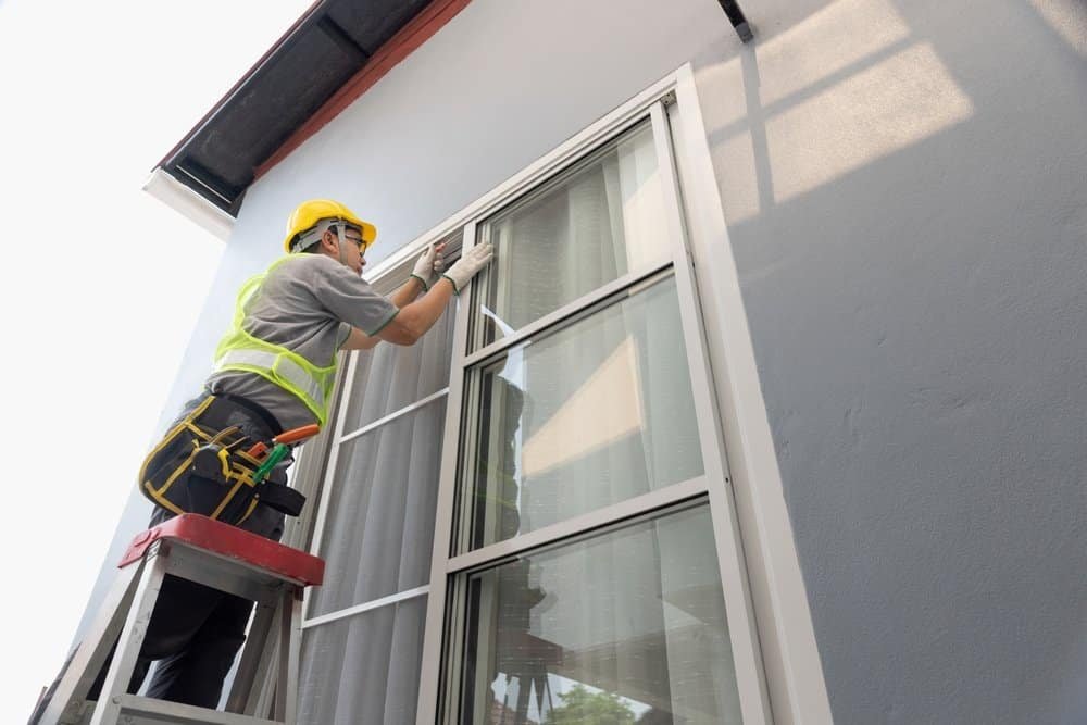 A worker wearing a yellow hard hat and safety vest stands on a ladder, installing or inspecting a window on the exterior of a house. Tools are attached to the worker's belt and the wall is light grey.