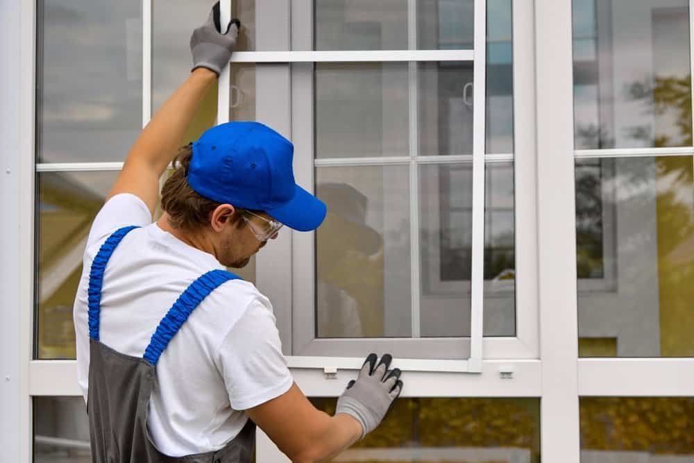 A worker in a blue cap, safety glasses, and gloves is installing or adjusting a window screen on a white-framed window.