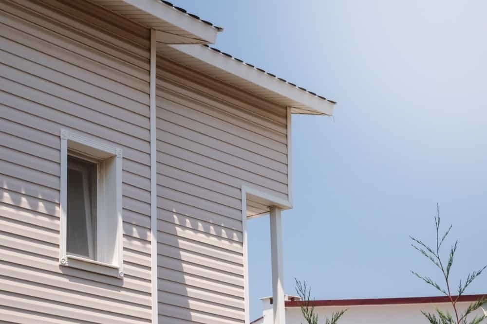 Close-up of the corner of a beige house with vinyl siding, a small white-framed window, and a sloped roof against a clear blue sky. Some thin branches are visible in the lower right.