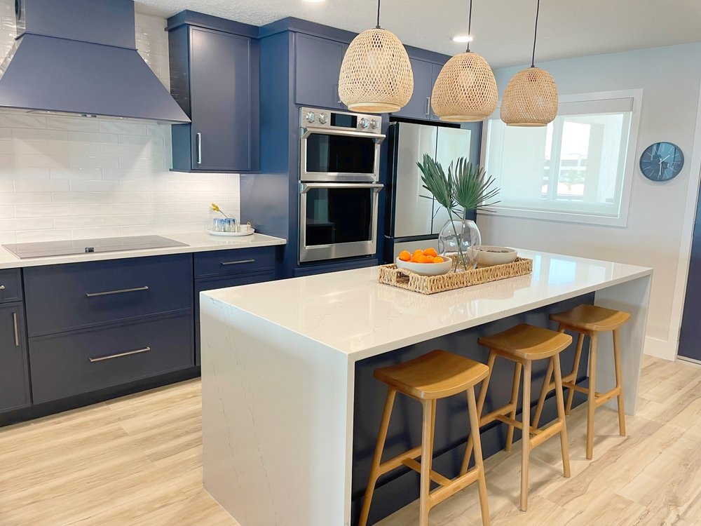 Modern kitchen with navy blue cabinets, stainless steel appliances, and a large white island featuring wooden stools. Woven pendant lights hang above the island, which holds a tray with dishes, fruit, and a leafy plant.