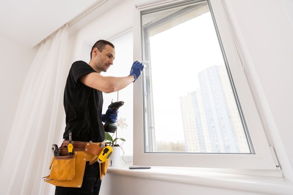 A person wearing a tool belt and blue gloves is fixing or cleaning an open window in a bright room, with tall apartment buildings visible outside.